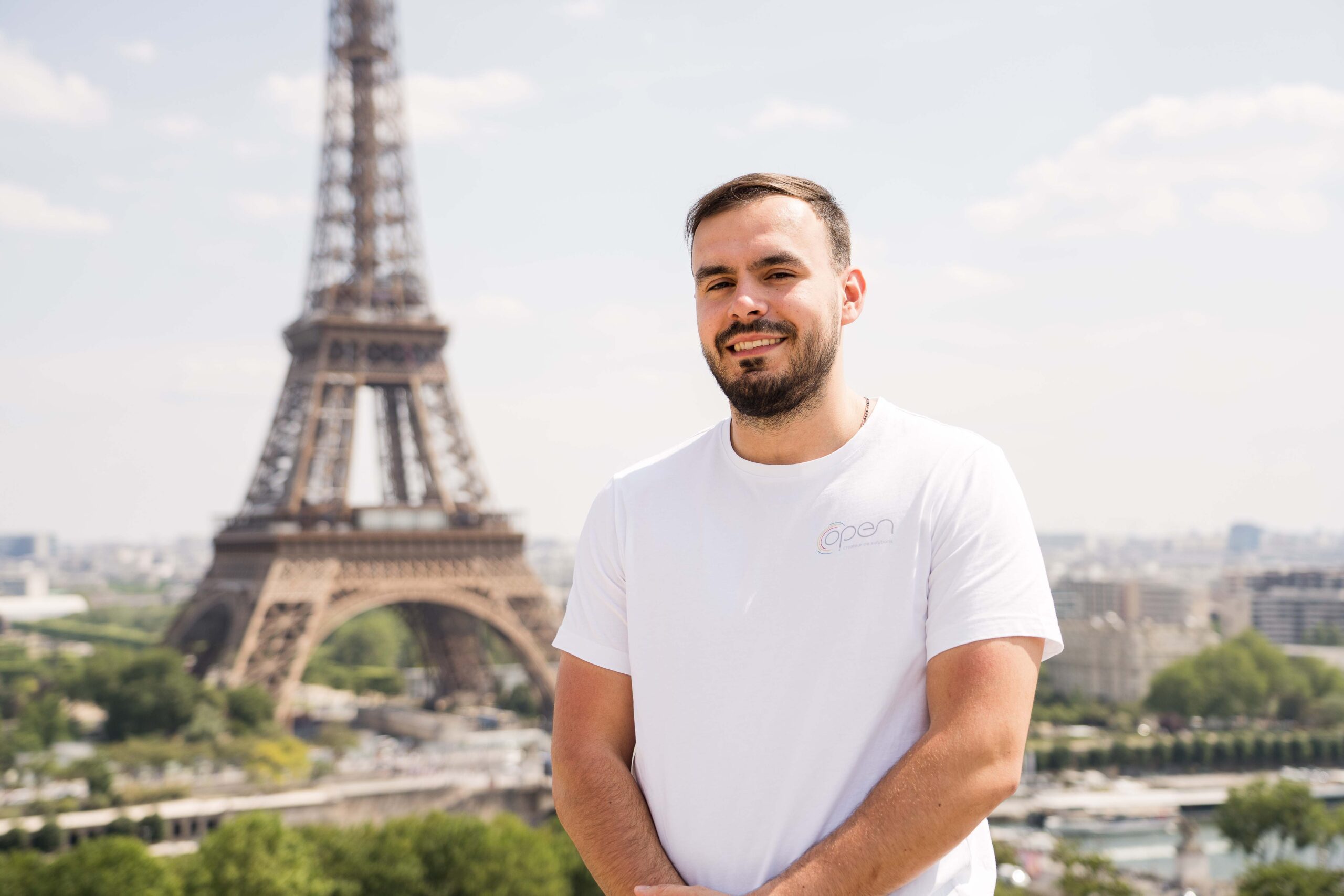 Un homme debout devant la tour Eiffel, vêtu d'un tee shirt blanche.