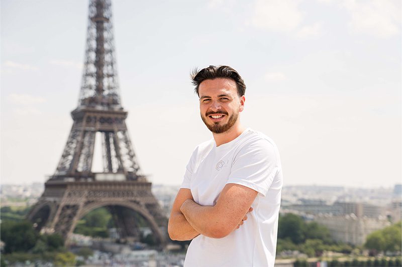 Un homme debout devant la tour Eiffel, vêtu d'un tee shirt blanc.