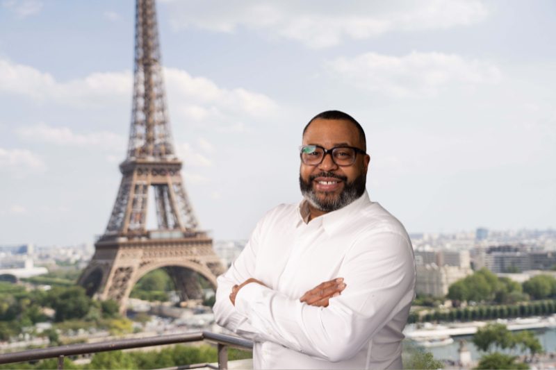 Un homme avec des lunettes et une chemise blanche debout devant la tour Eiffel.